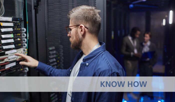 Ein Techniker in blauem Hemd richtet im Rechenzentrum ein Serverrack ein, im Hintergrund sind Kollegen zu sehen. LED-Leuchten beleuchten den Serverraum und schaffen eine moderne, technologische Atmosphäre.